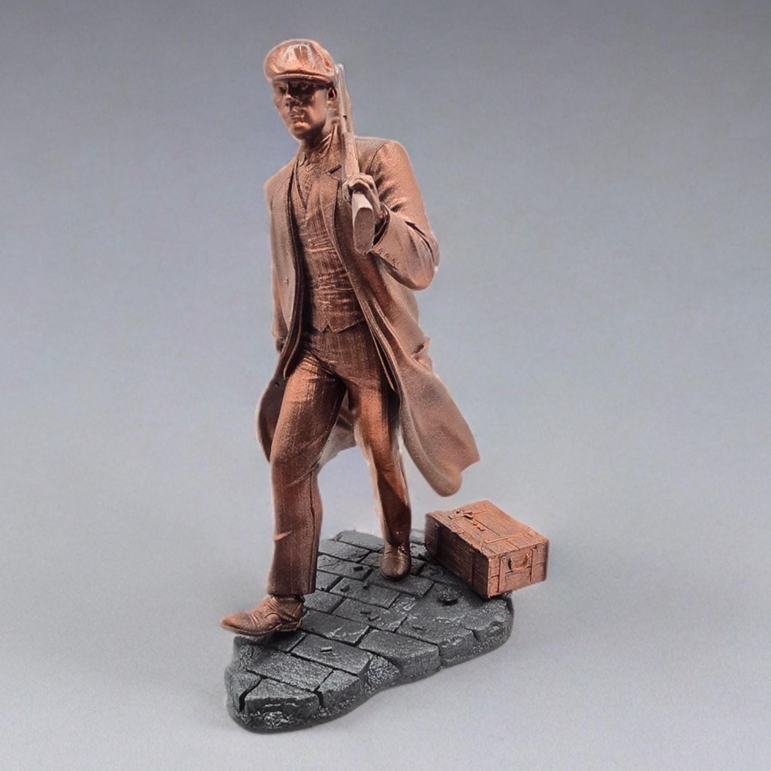 Bronze-colored 3D-printed figurine of a man in a long coat and newsboy cap, carrying a rifle. He stands on a dark gray, textured base resembling cobblestones. The figurine is detailed, showing the man's clothing and the rifle's features.  The overall style suggests a gangster or historical figure theme. The figurine is a collectible item, suitable for display.