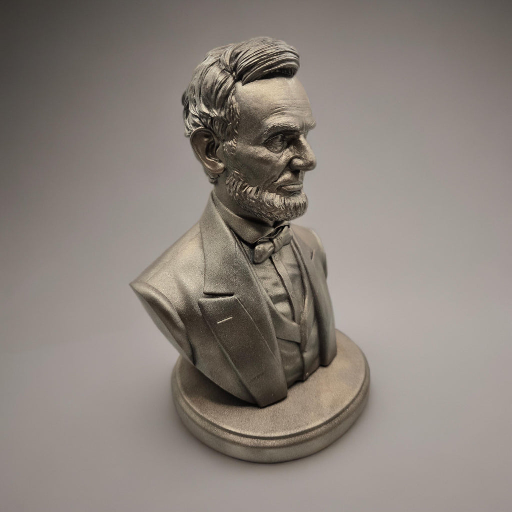 Silver-colored bust statuette of Abraham Lincoln, showcasing detailed facial features, a neatly trimmed beard, and formal attire with a bow tie, mounted on a rectangular base.