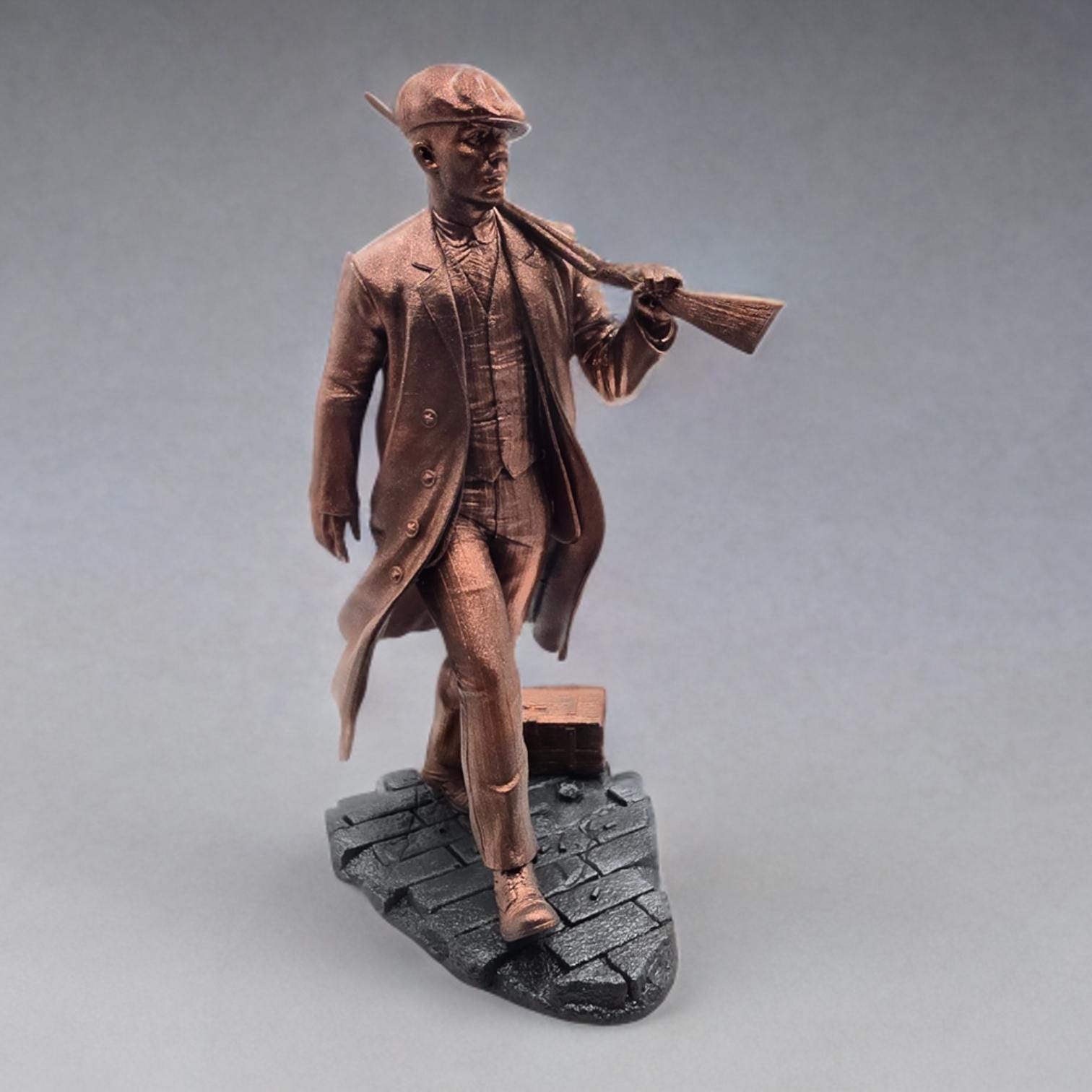 Bronze-colored 3D-printed figurine of a man in a long coat and newsboy cap, carrying a rifle. He stands on a dark gray, textured base resembling cobblestones. The figurine is detailed, showing the man&#39;s clothing and the rifle&#39;s features.  The overall style suggests a gangster or historical figure theme. The figurine is a collectible item, suitable for display.