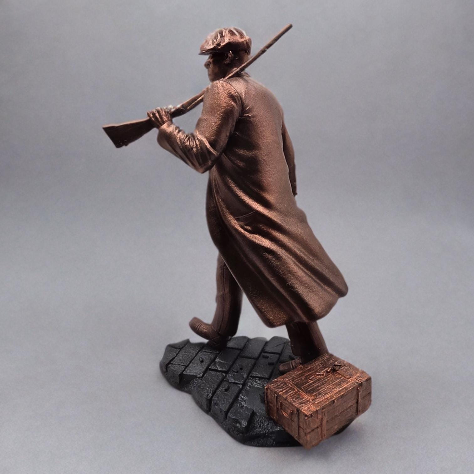 Bronze-colored 3D-printed figurine of a man in a long coat and newsboy cap, carrying a rifle. He stands on a dark gray, textured base resembling cobblestones. The figurine is detailed, showing the man's clothing and the rifle's features.  The overall style suggests a gangster or historical figure theme. The figurine is a collectible item, suitable for display.