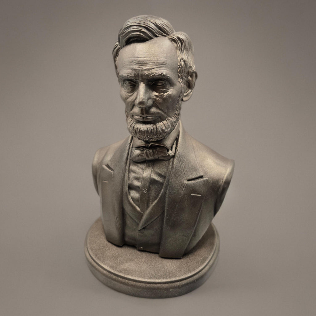Silver-colored bust statuette of Abraham Lincoln, showcasing detailed facial features, a neatly trimmed beard, and formal attire with a bow tie, mounted on a rectangular base.