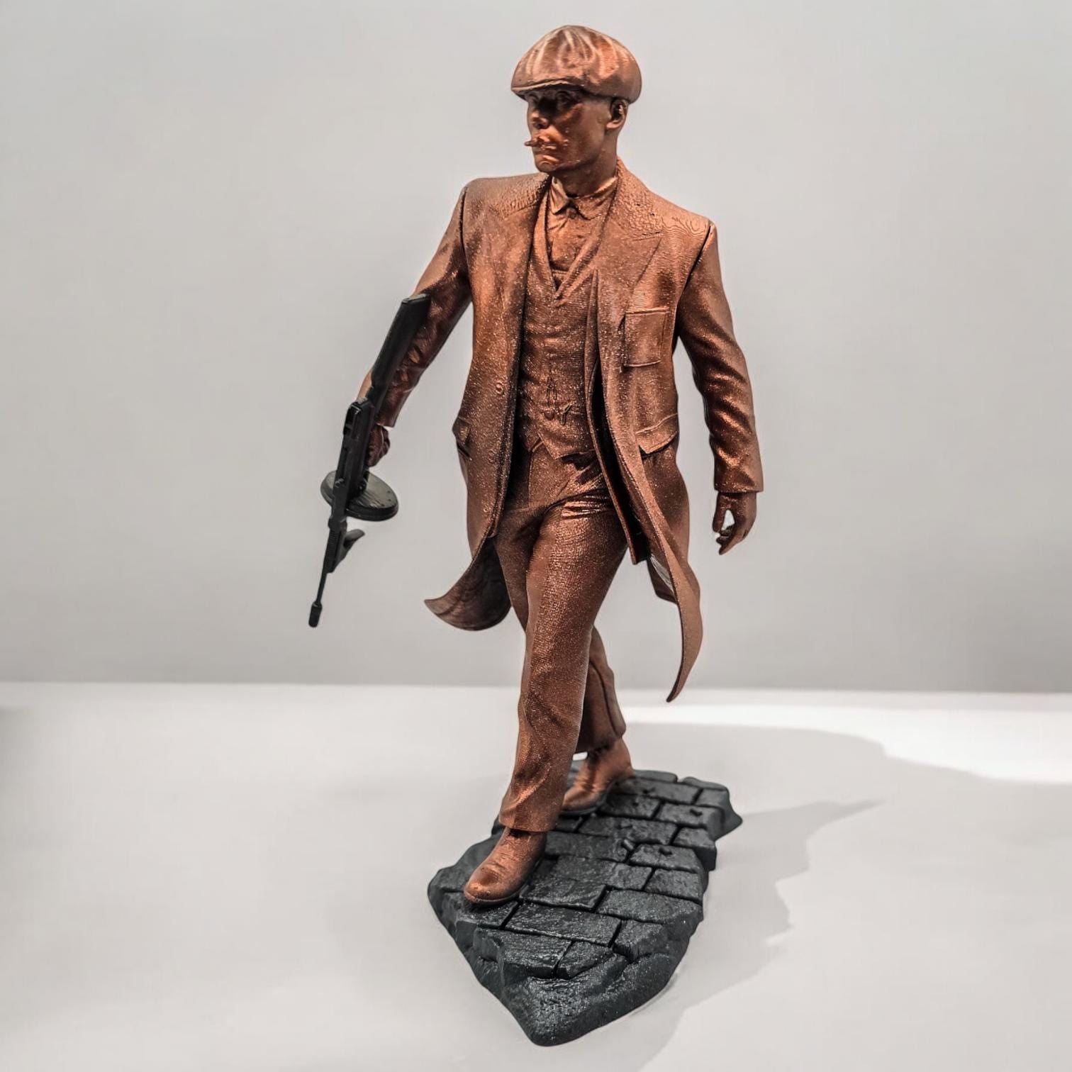 A bronze-colored figurine depicts a man in a long coat and newsboy cap, holding a submachine gun. He stands on a textured, dark gray base, styled as a cobblestone street. The figurine is detailed, capturing the texture of the fabric and the weapon.  The overall style suggests a gangster or 1920s era figure.