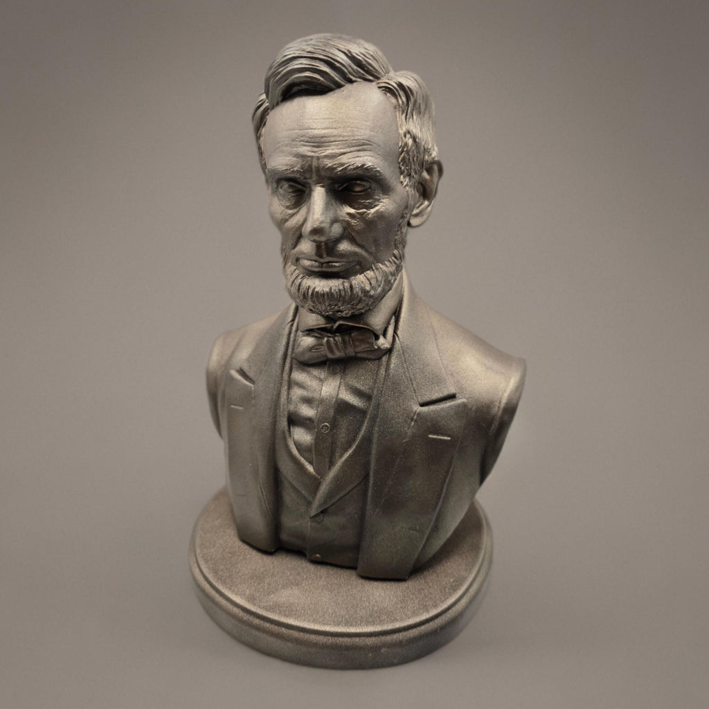 Silver-colored bust statuette of Abraham Lincoln, showcasing detailed facial features, a neatly trimmed beard, and formal attire with a bow tie, mounted on a rectangular base.