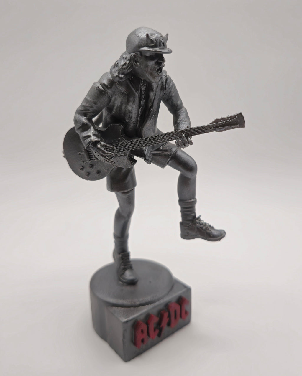 Statue of Angus Young from AC DC, depicted in a dynamic pose with his iconic guitar and schoolboy uniform, standing on a base engraved with the band&#39;s logo.