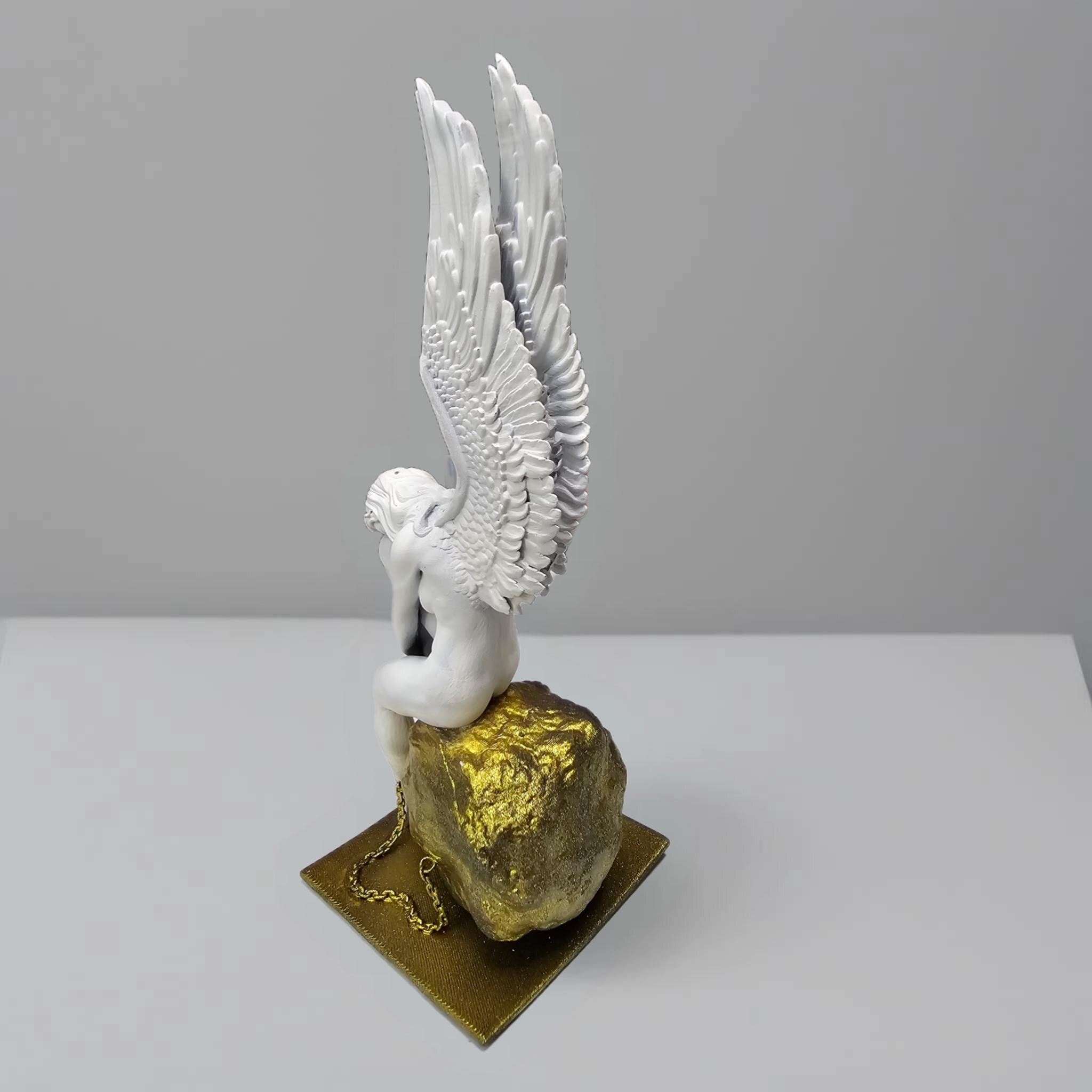 Statue of a chained angel seated on a textured pedestal, with detailed wings and a contemplative pose.