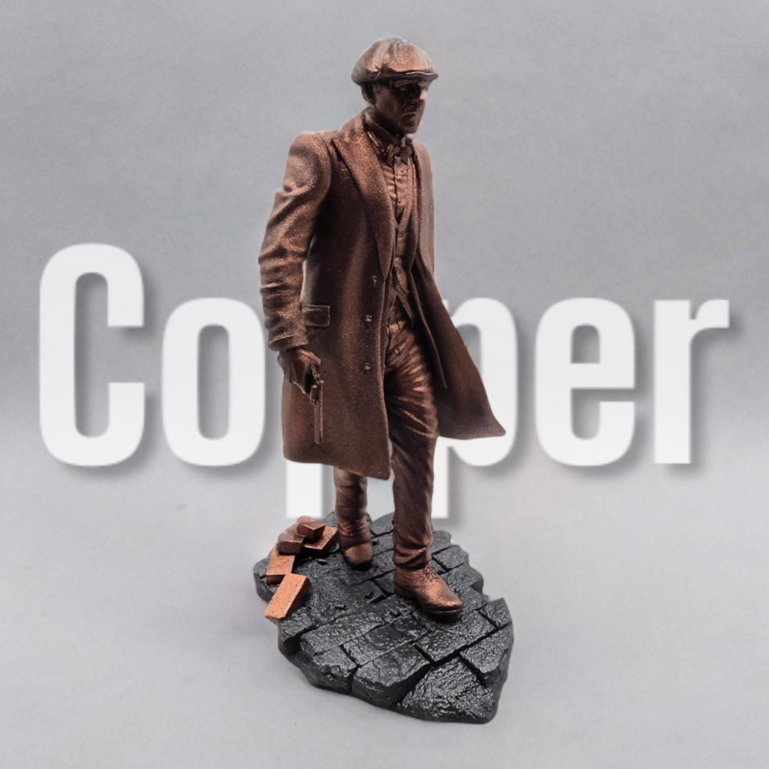 A bronze-colored figurine depicts a man in a long coat and newsboy cap, holding a submachine gun. He stands on a textured, dark gray base, styled as a cobblestone street. The figurine is detailed, capturing the texture of the fabric and the weapon.  The overall style suggests a gangster or 1920s era figure.